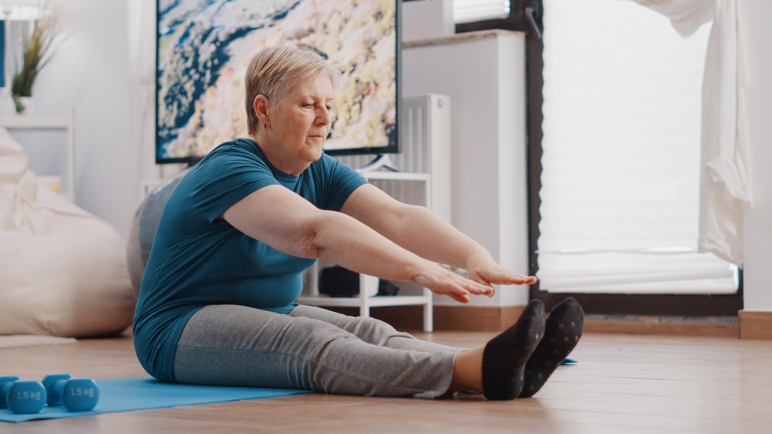 Aged woman doing stretching exercise on yoga mat. Senior person bending over to stretch arms and legs muscles at home for wellness and fitness. Elder adult exercising and training