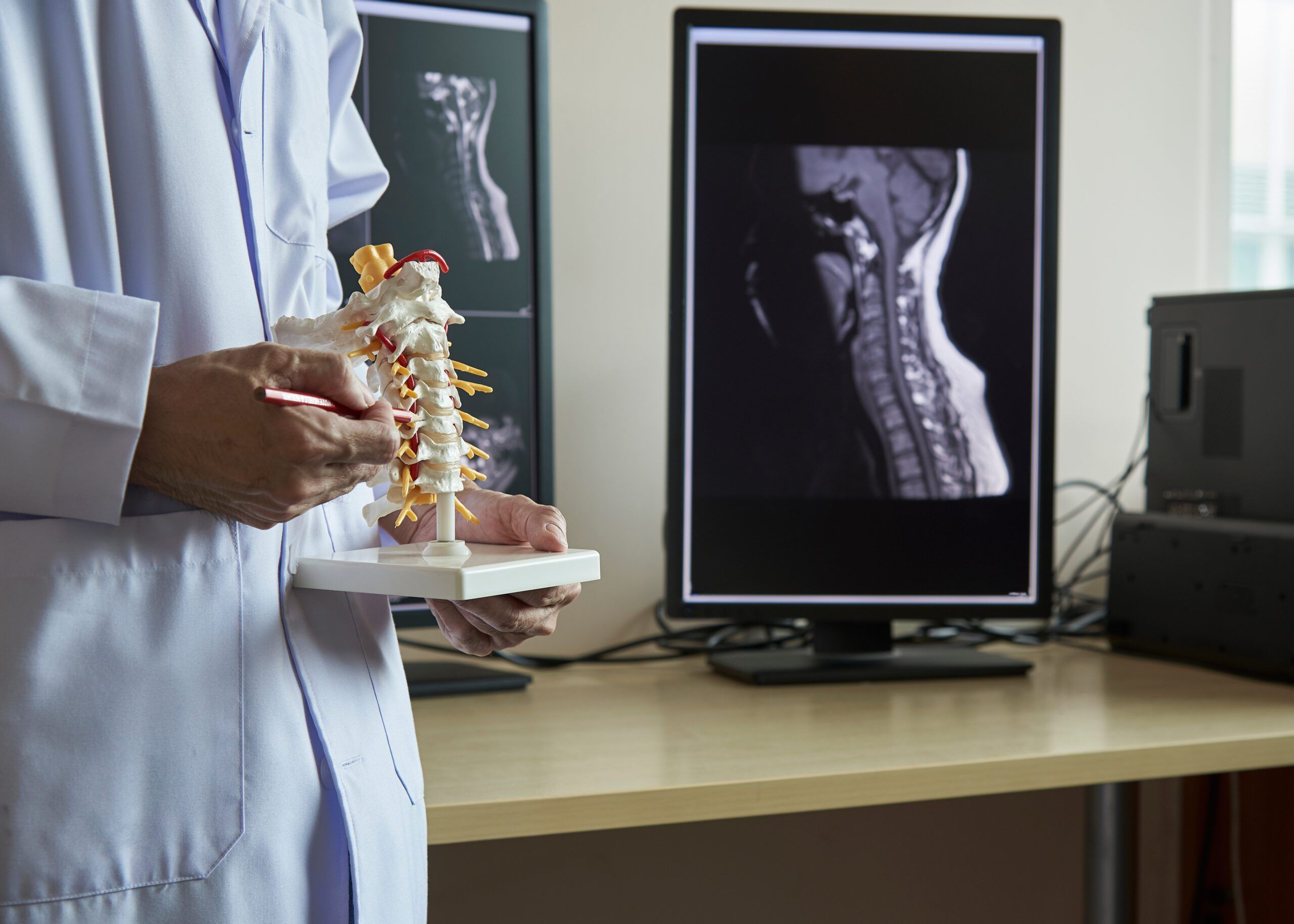 A neurosurgeon pointing at anatomy of human cervical spine model in medical office. Lumbar spine x-ray in computer screen on background.